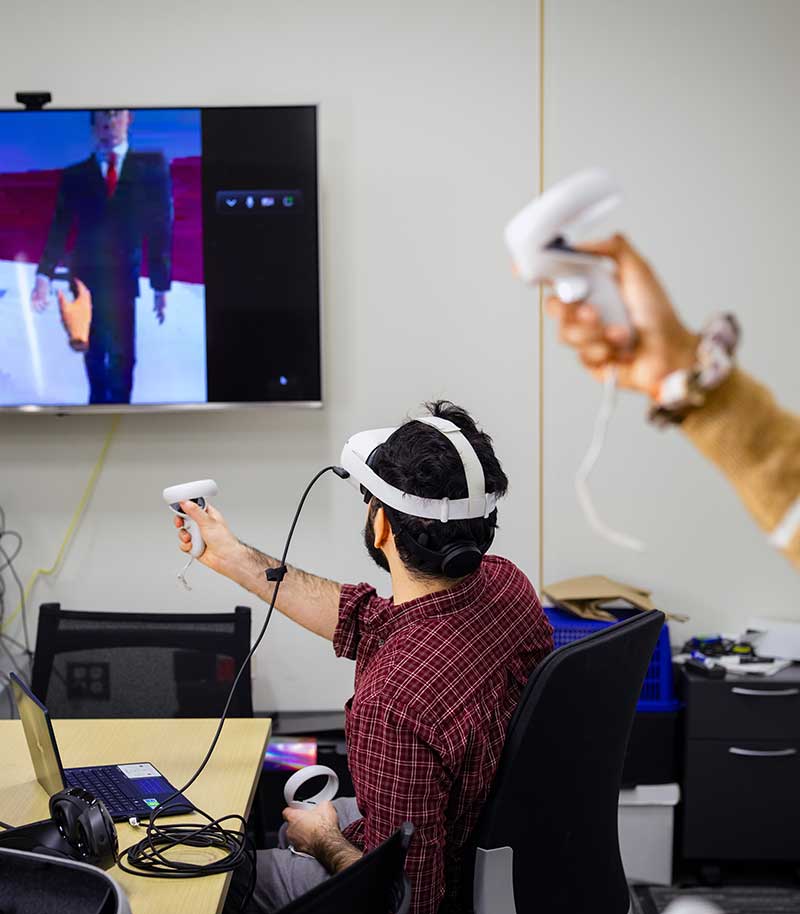 A student wearing a virtual reality headset and holding a game controller in one hand sits at a large table in a conference room. Another student holds a controller while standing behind the first student. A game is displayed on a large TV screen hanging on the wall of the room.