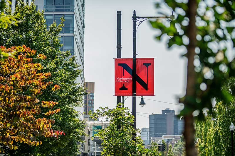 A red banner hanging from a streetlight pole. The banner shows Northeastern's black N wordmark.