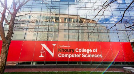 A view of the glass exterior of West Village H with a large, red Khoury College of Computer Sciences banner hanging across several windows.