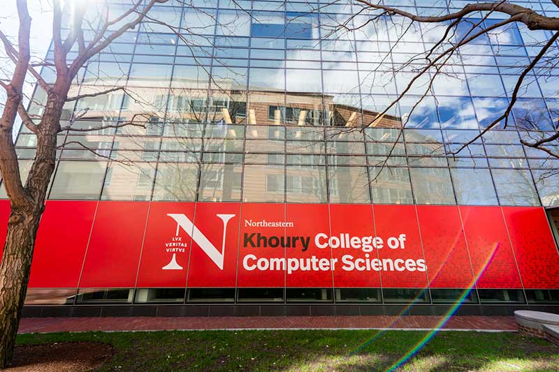 A view of the glass exterior of West Village H with a large, red Khoury College of Computer Sciences banner hanging across several windows.