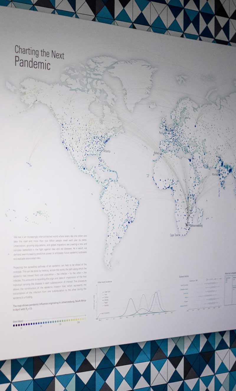 A large poster titled 'Charting the Next Pandemic' hangs on a wall in the Network Science lab. The poster has a map of the world with many blue and purple dots marking different cities.