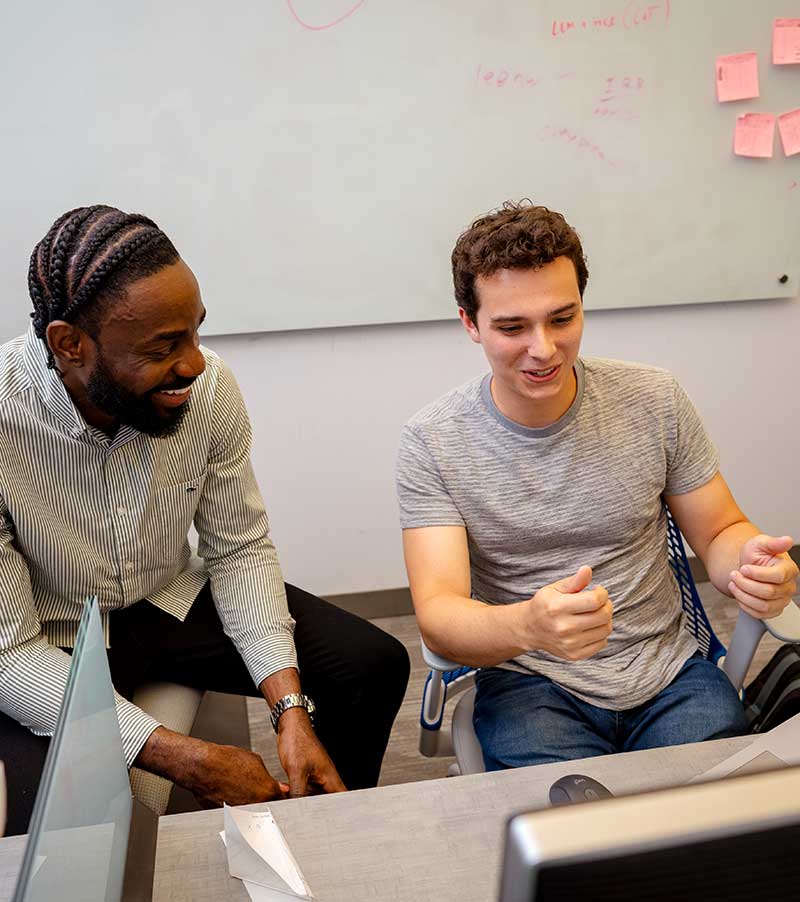 Two students sit at a desk in front of a computer. The student on the right is gesturing toward the screen with his hands.