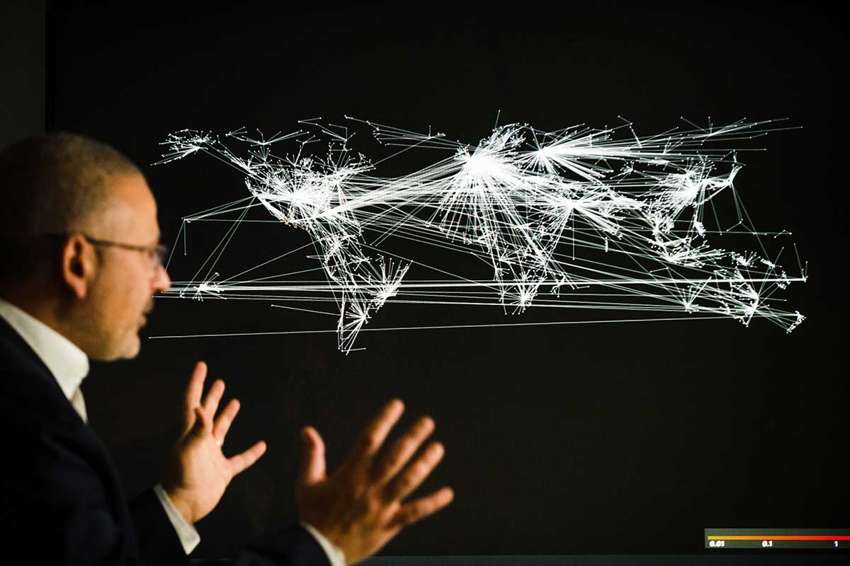 Khoury faculty member Alessandro Vespignani stands in front of a screen showing a black and white map of the world with many white lines conneecting different cities on the map.