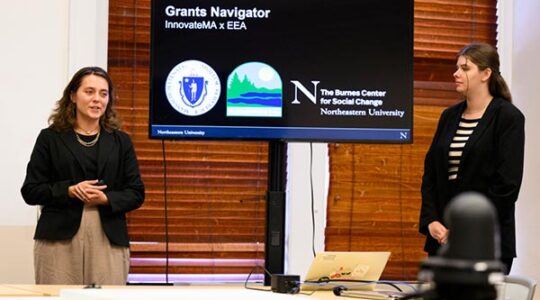 Two Khoury students stand alongside a large screen that shows their Grants Navigator presentation