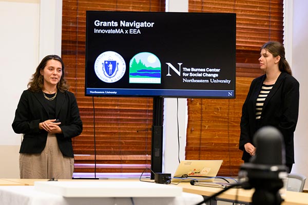 Two Khoury students stand alongside a large screen that shows their Grants Navigator presentation