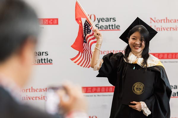 A Northeastern graduate holds small flags of the United States and China in one hand and her diploma in the other hand.