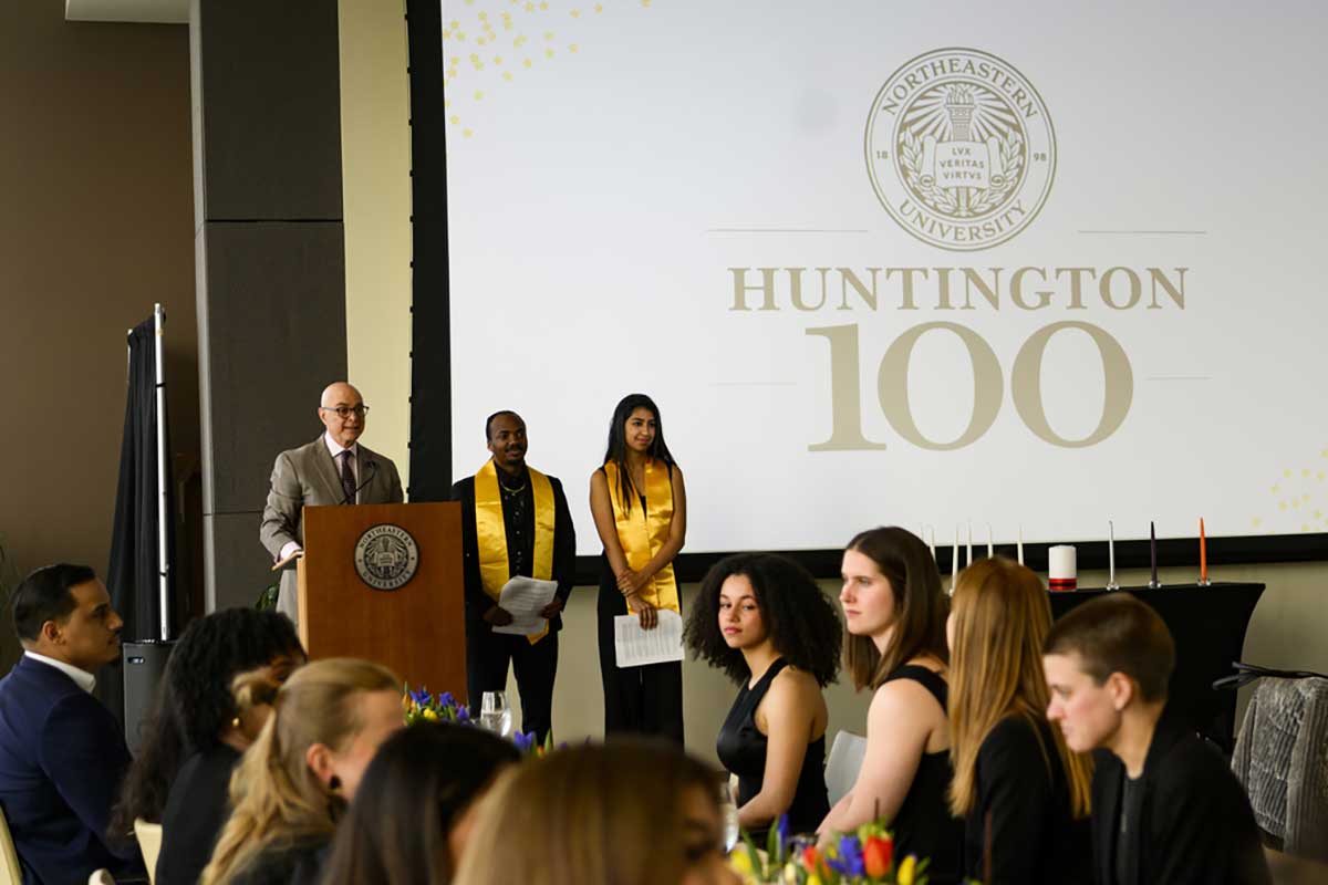 President Joseph Aoun (left) speaks at a podium while two members of the Huntington 100 stand at right.