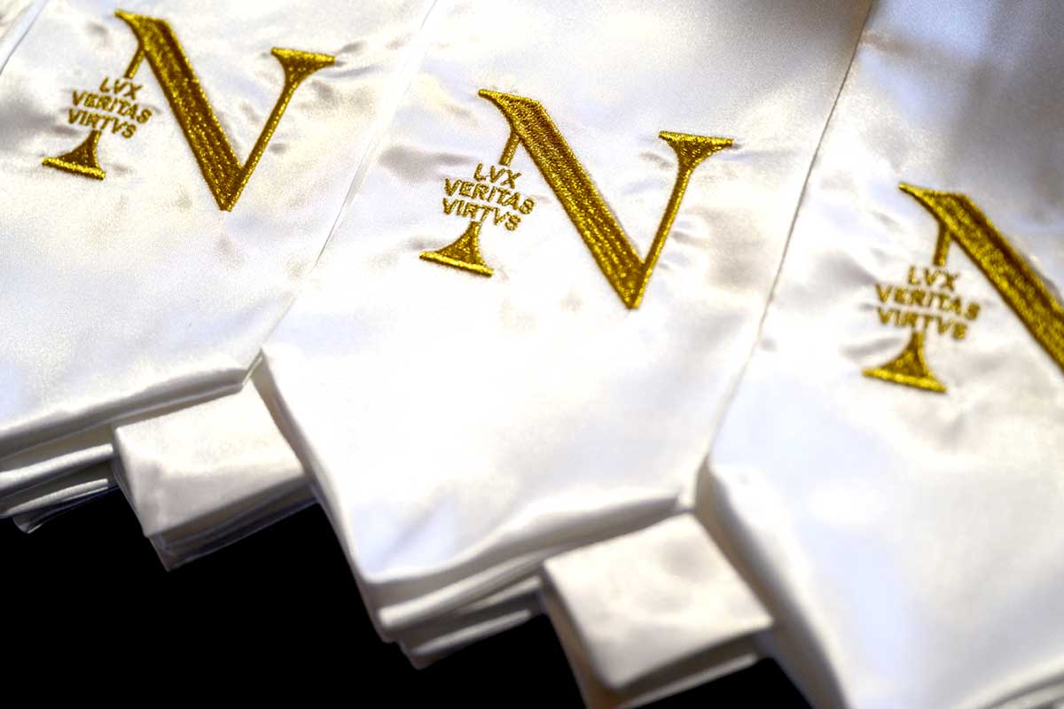 Several white graduation stoles lay on a table. The stoles have the Northeastern N logo embroidered in gold fabric.