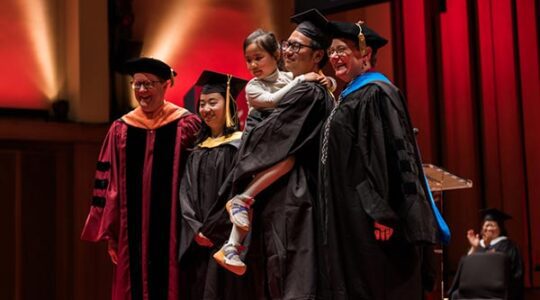 Two Northeastern graduates stand on a stage with two members of Northeastern administration. One graduate is holding a small child in his arms.