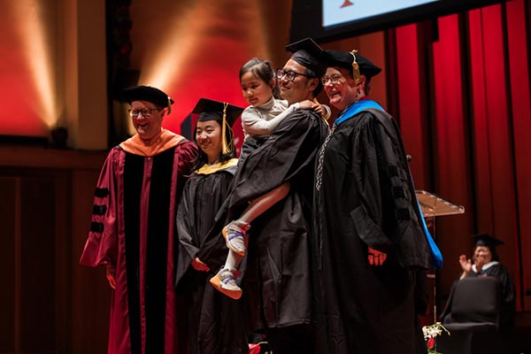 Two Northeastern graduates stand on a stage with two members of Northeastern administration. One graduate is holding a small child in his arms.