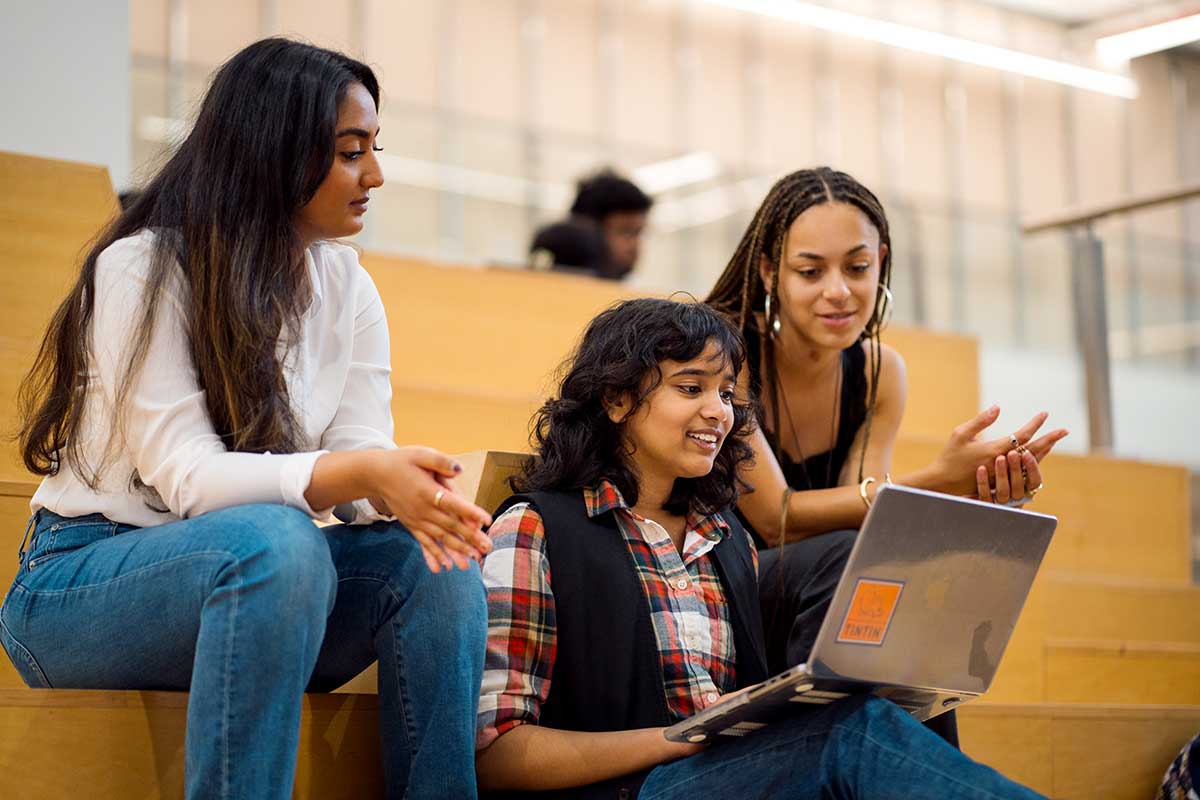 Three students sitting on ampitheater style benches discuss a project. The student in the middle has a laptop open and resting on her knees.