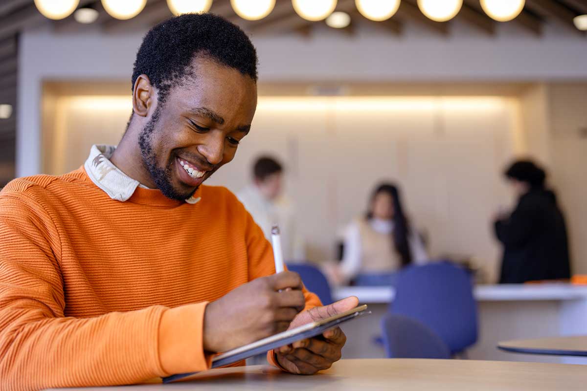 A student sitting at a large table smiles as he writes in a notebook