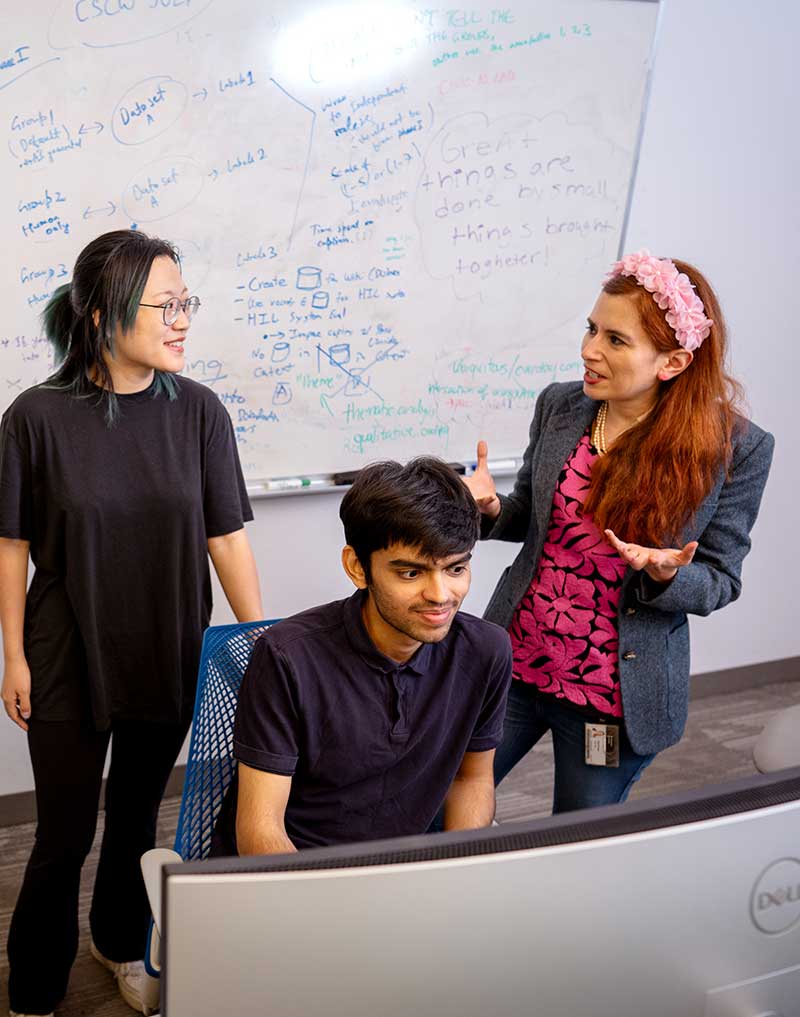 Khoury professor Saiph Savage, right, discusses research with two students in a Khoury lab. One student is sitting at a desk and the other is standing in front of a whiteboard filled with writing.