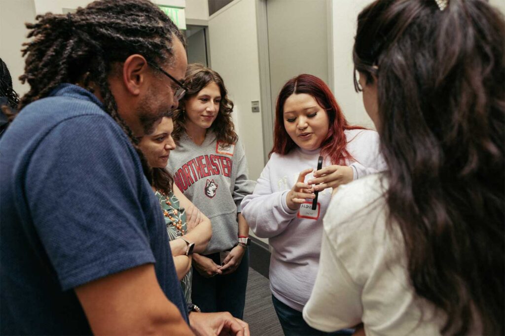 A Microsoft holds a mobile device and demonstrates an app's functionality while Khoury interns look on