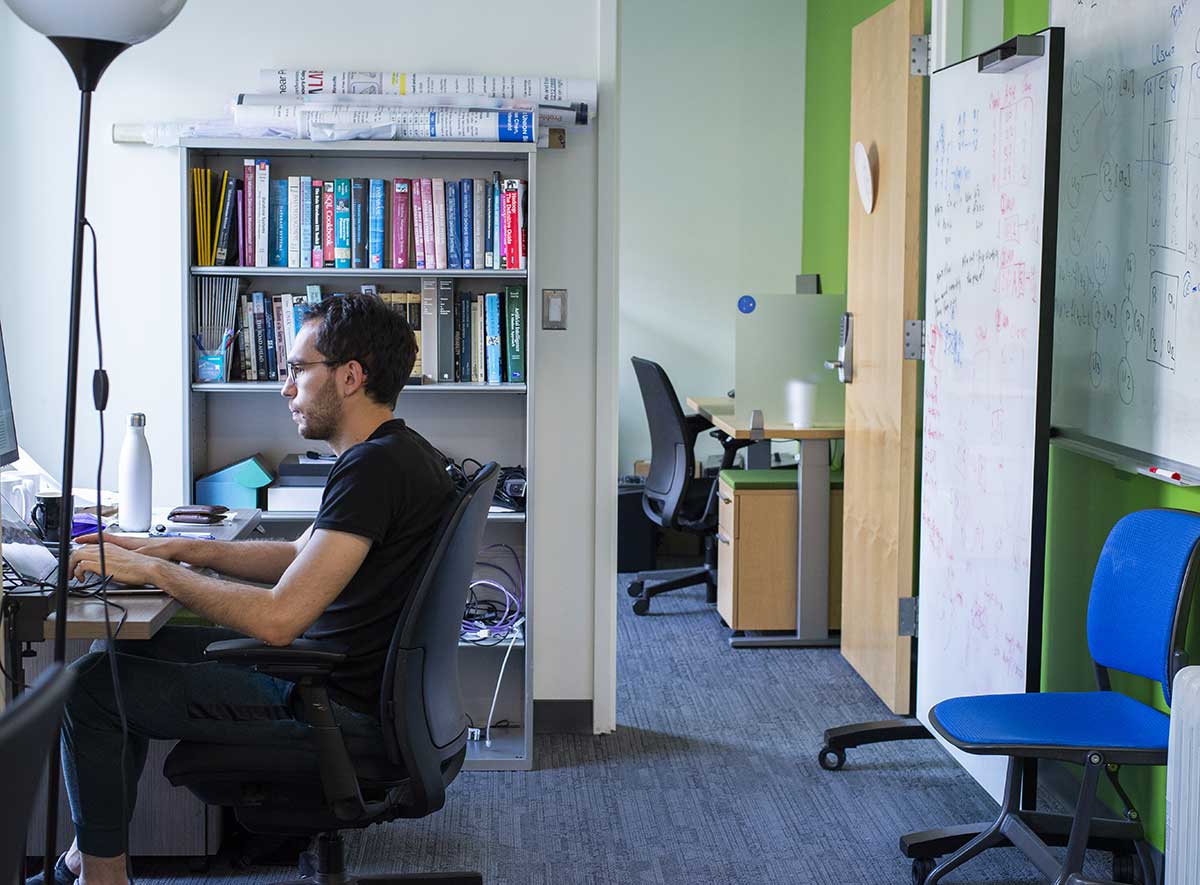 A Khoury data management student works types on a computer keyboard in a college lab. A whiteboard filled with mathematical equations and notes is behind the student.