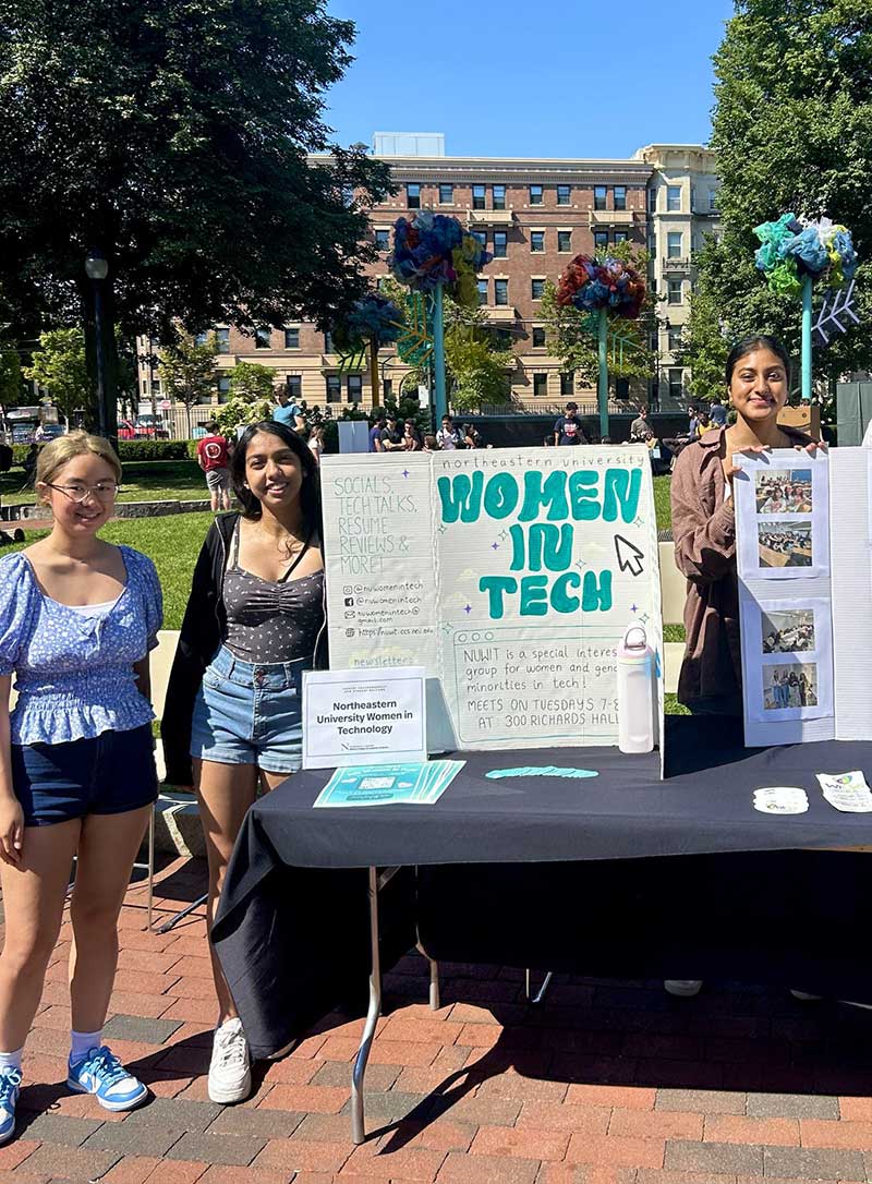 Three members of Northeastern's Women In Tech student club stand around their poster during a club fair at Northeastern