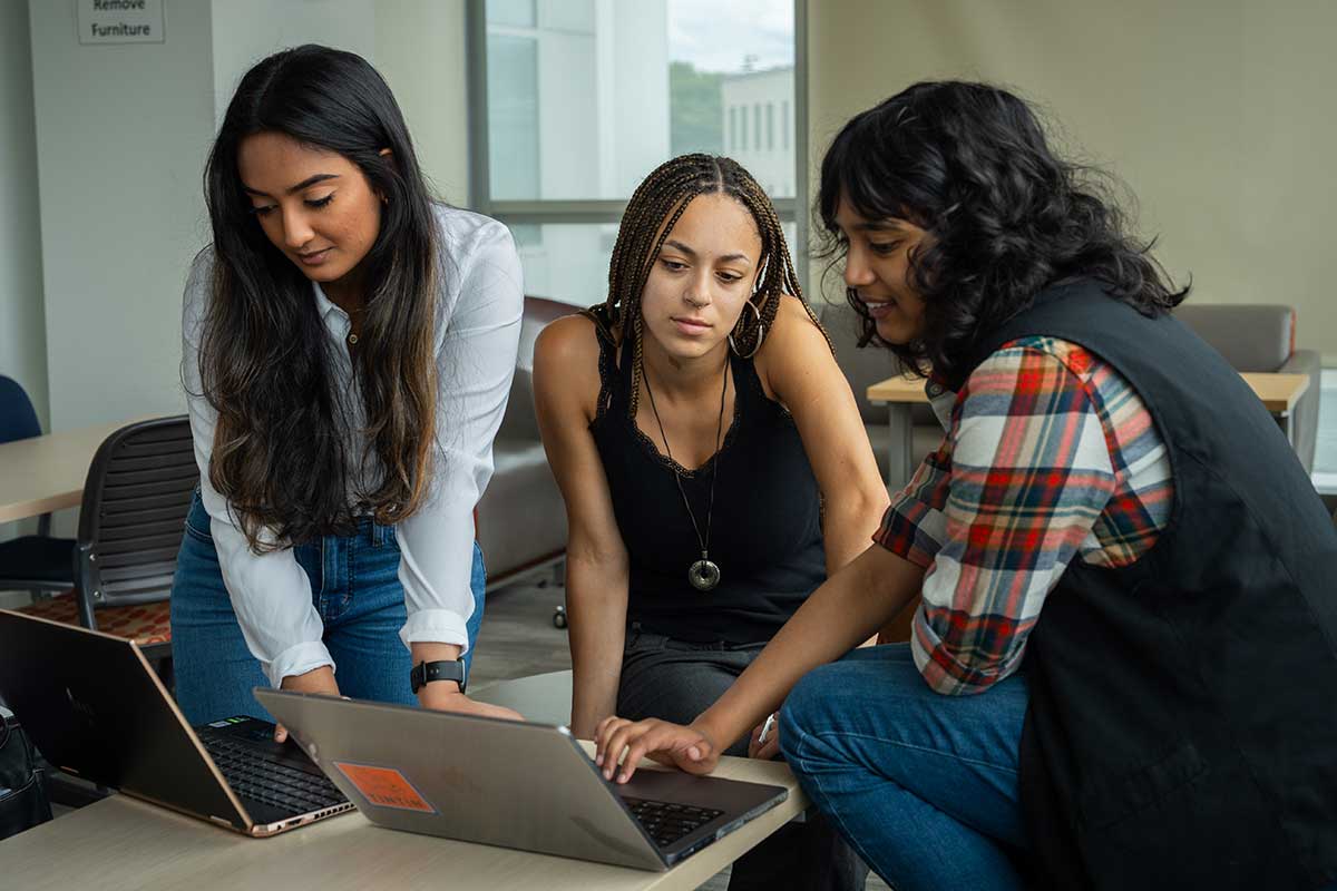 A group of three students sit around two open laptops and work on a project in a Northeastern lounge