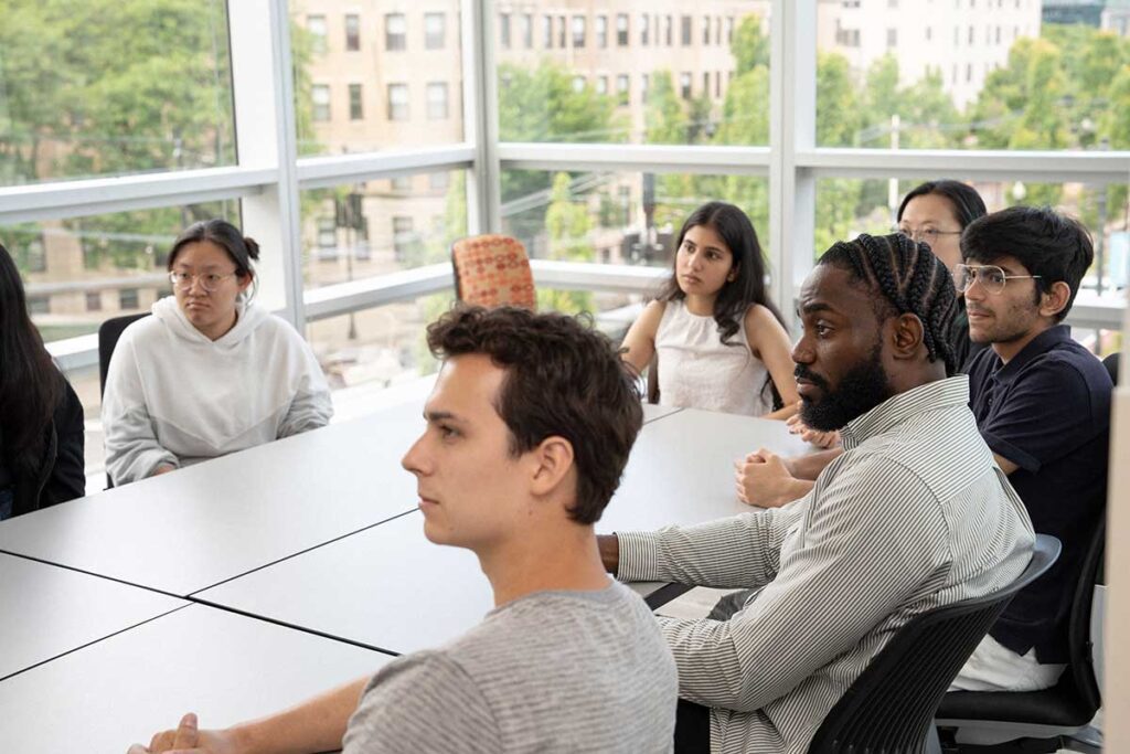 Six students sit around a large rectangular table in a West Village H classroom and watch a presentation