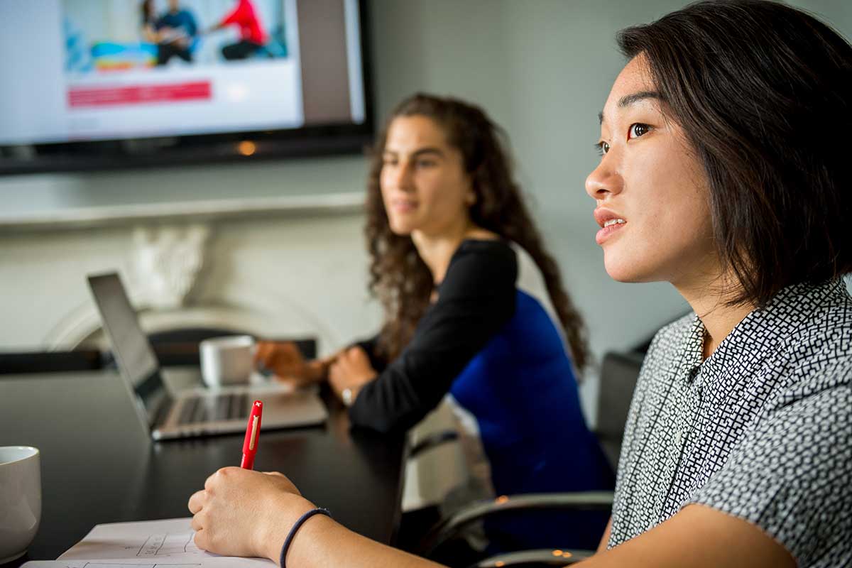 A Khoury student sits at a conference table with an employee at a co-op partner at left. There is large screen showing a photo hanging on the wall.