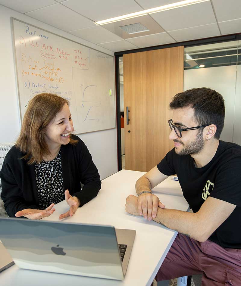 A Khoury faculty members sits to the left of a table speaking with a student sitting to the right of the table. An open laptop sits on the desk in front of them.