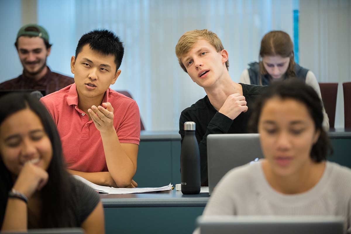 Three rows of students view a course lecture. The student in the middle of the photo is asking a question.