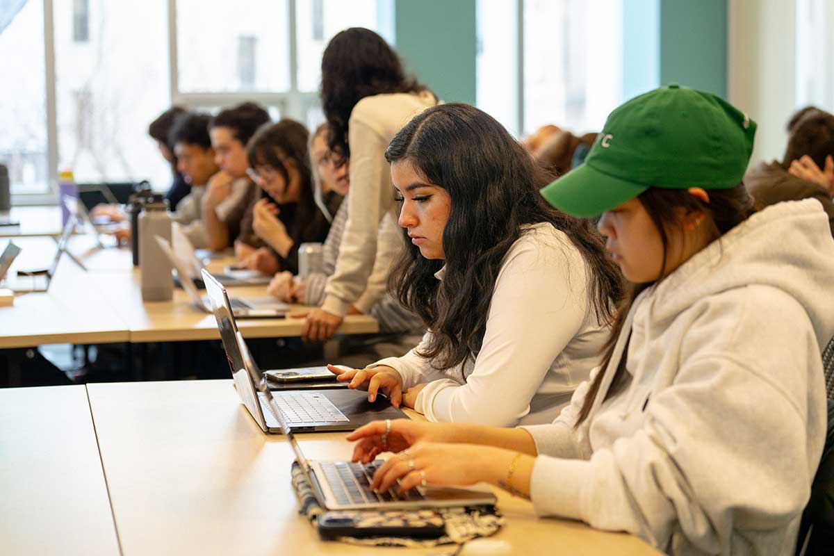 Several students sitting at large, rectangular tables work during a class