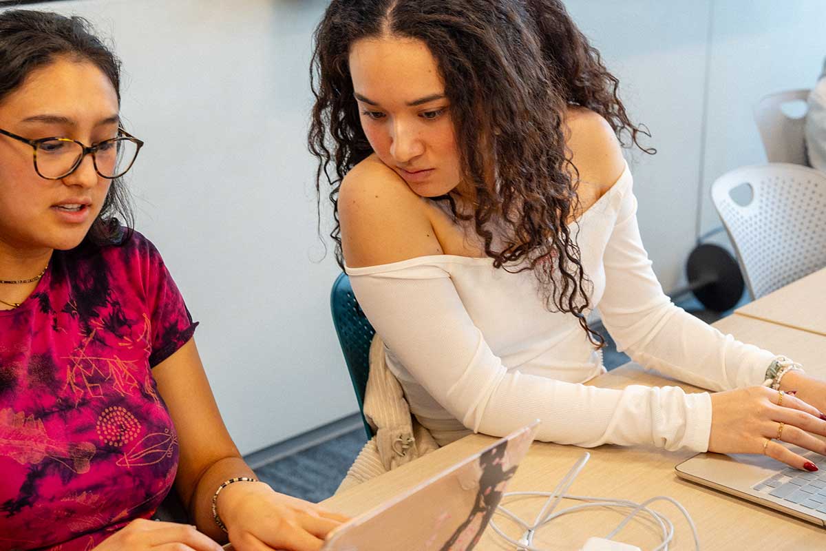 Two students sitting at a large table view an open laptop. The student on the right types on a different laptop.