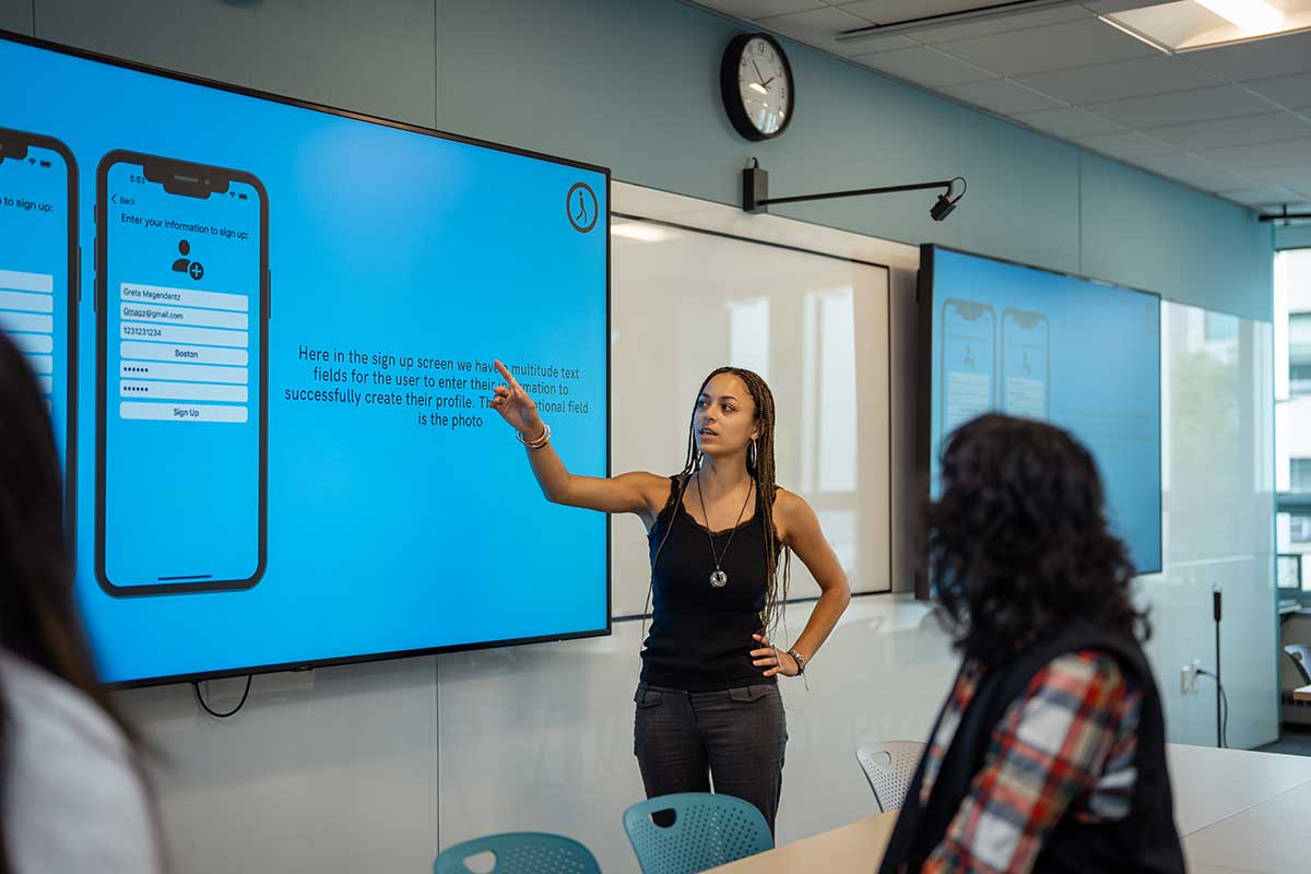 A Khoury student points at a projection screen that's showing two mobile devices with an information form displayed.