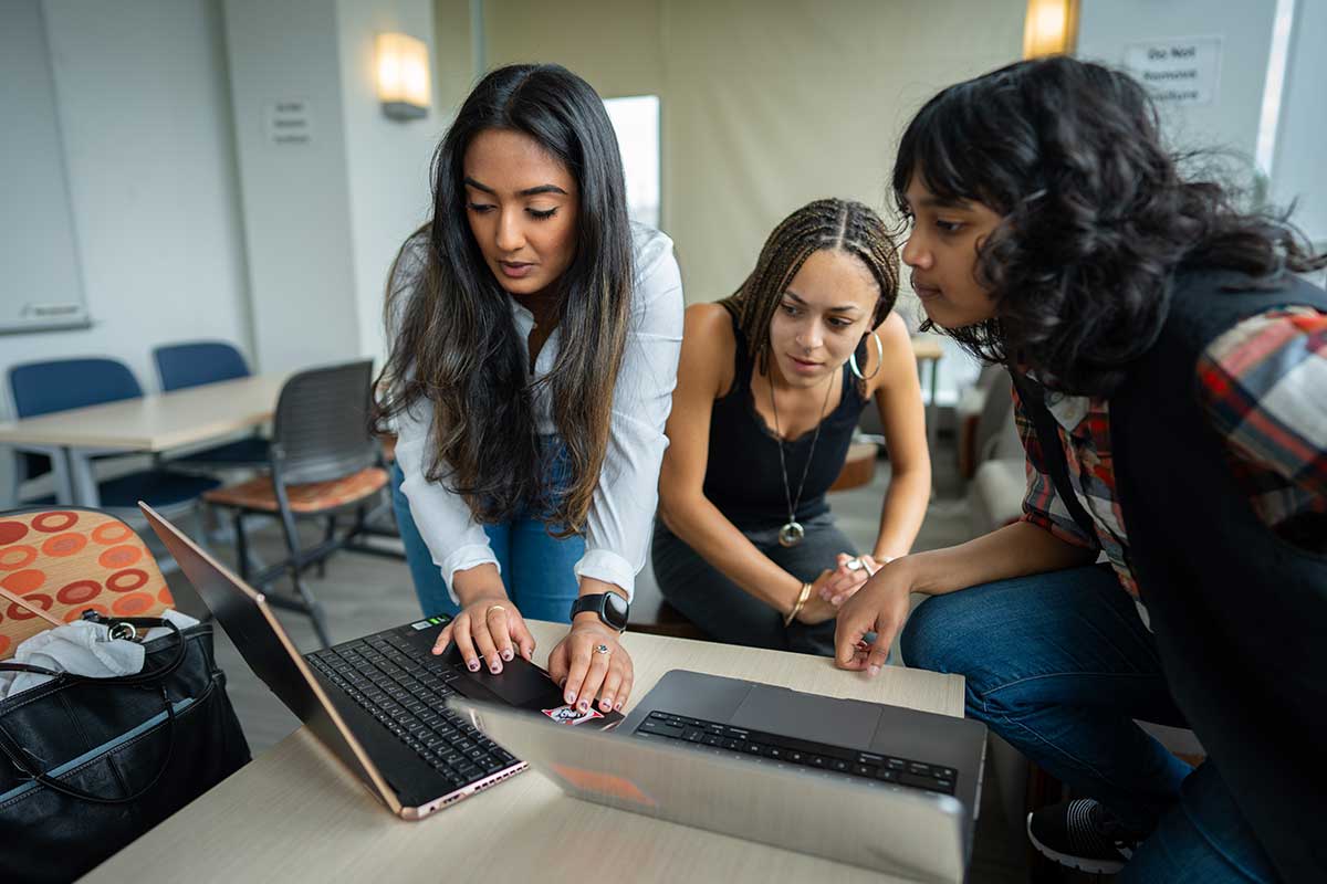 Three students view an open laptop on a table