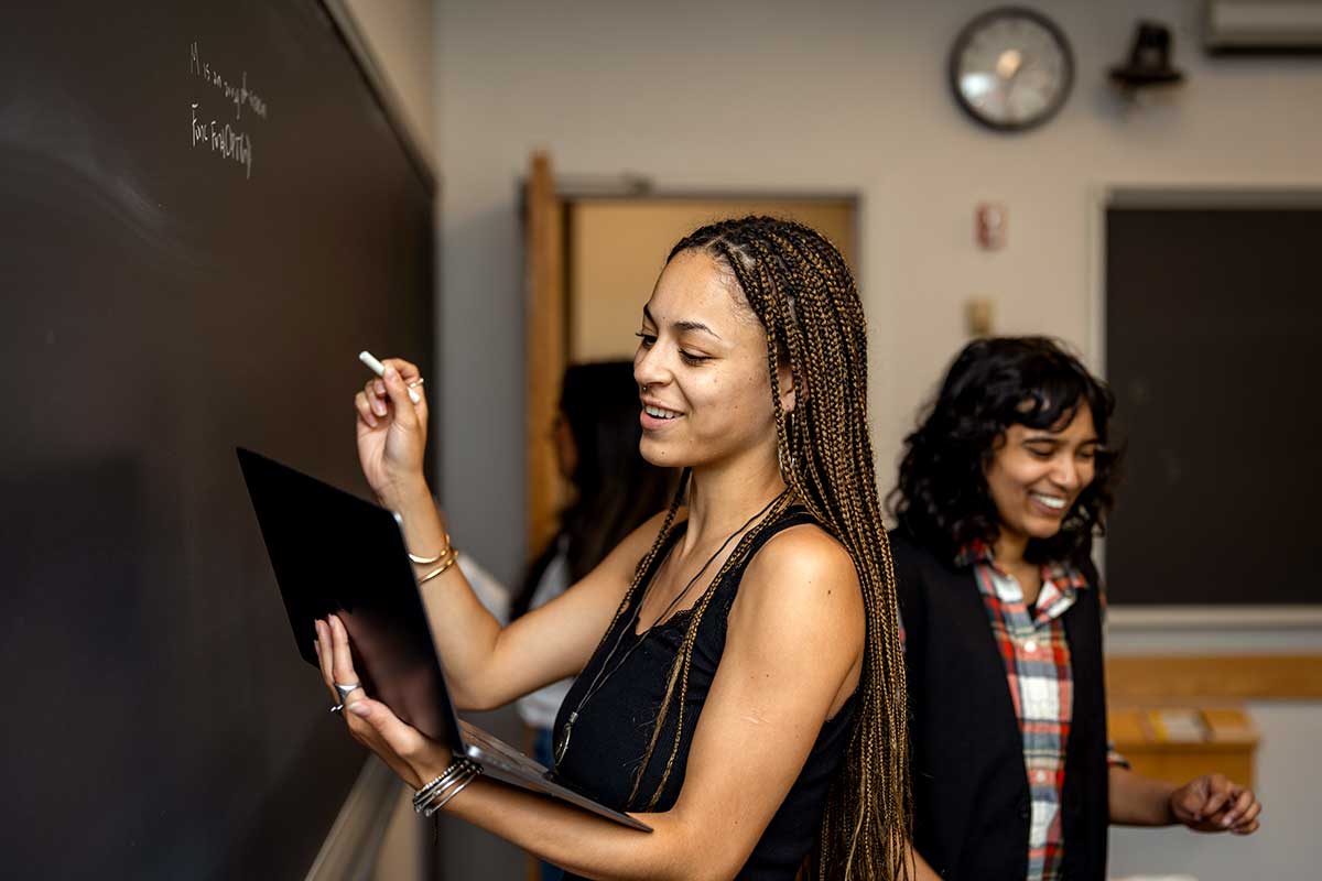 A student writes on a chalkboard while holding an open laptop in one arm while another student smiles