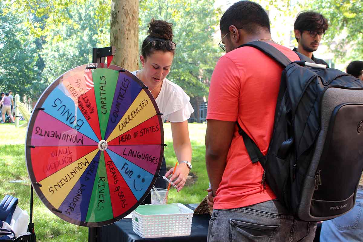 At a club fair, a student standing behind a table holding a gameshow-style wheel talks with another student