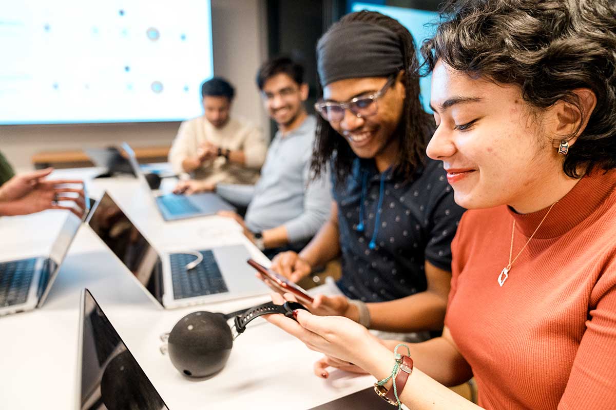 Four students sitting at a large table work on their laptops. In the foreground, a student holds a smart watch.