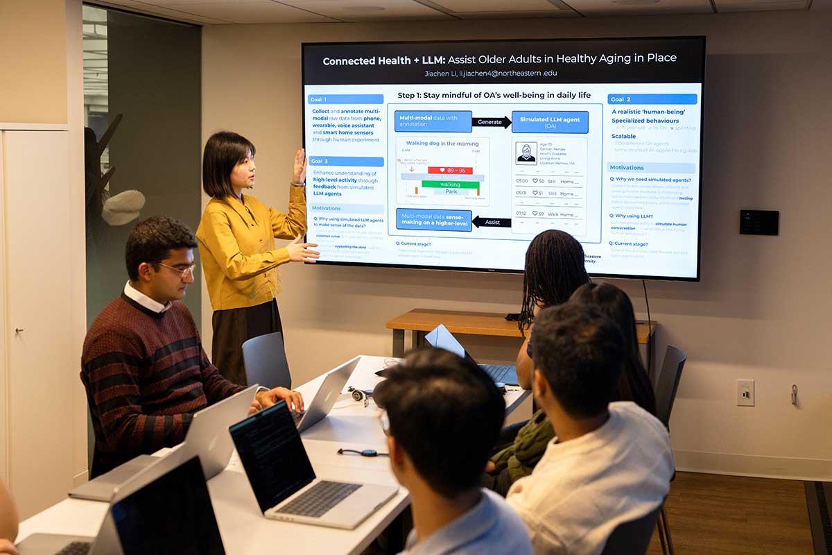 A gives a presentation to several Khoury faculty and students in a conference room. A large screen on the wall shows the presentation title: Connected Health + LLM: Assist Older Adults in Healthy Aging in Place