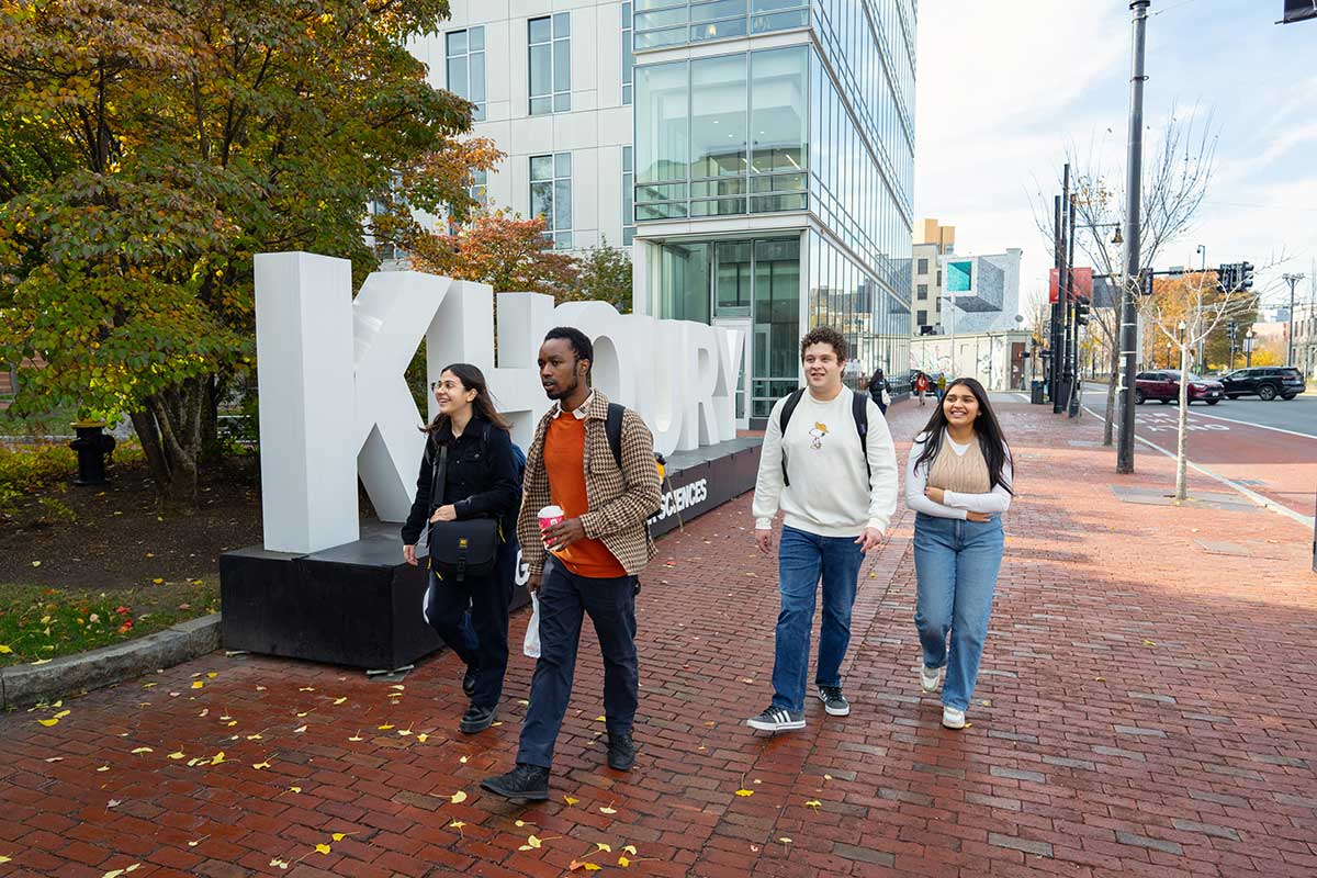 A group of 4 students talk as they walk on a brick sidewalk on Huntington Avenue in Boston