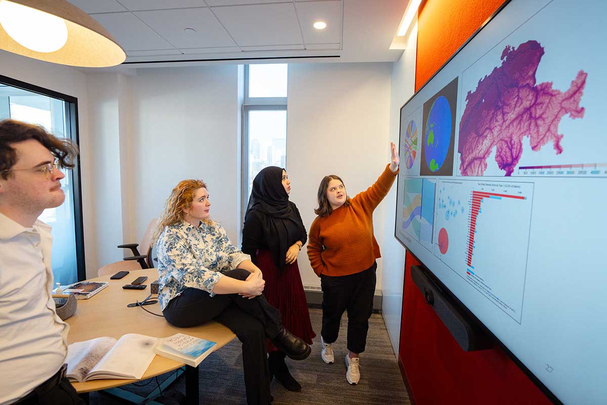 A Khoury PhD student gestures at a projection screen showing an image of the Earth and a bar chart while three other students look on