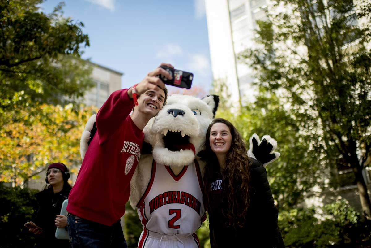 A student takes a selfie with Northeastern's husky mascot and another student.