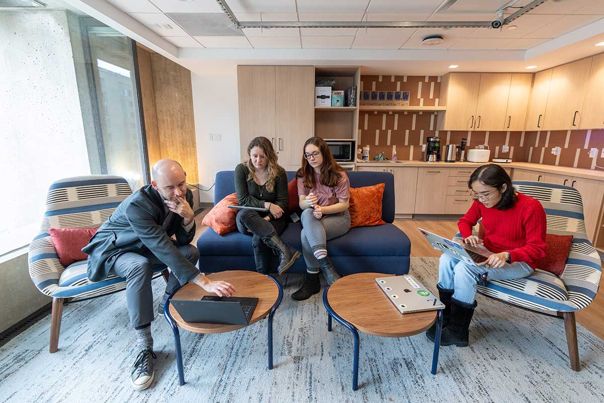Khoury faculty member Christo Wilson (left) types on a laptop while two students sitting next to him look on. Another student is sitting on a chair in the same lab.