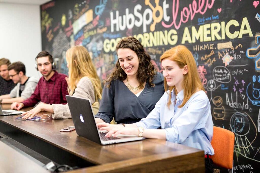 A student sitting at a large table types on a laptop while her co-op supervisor talks to her