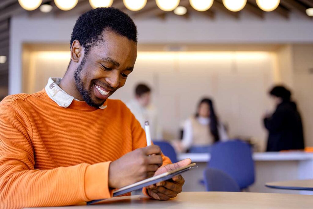 A student sits at a table in a campus lounge and writes with a stylus on a tablet