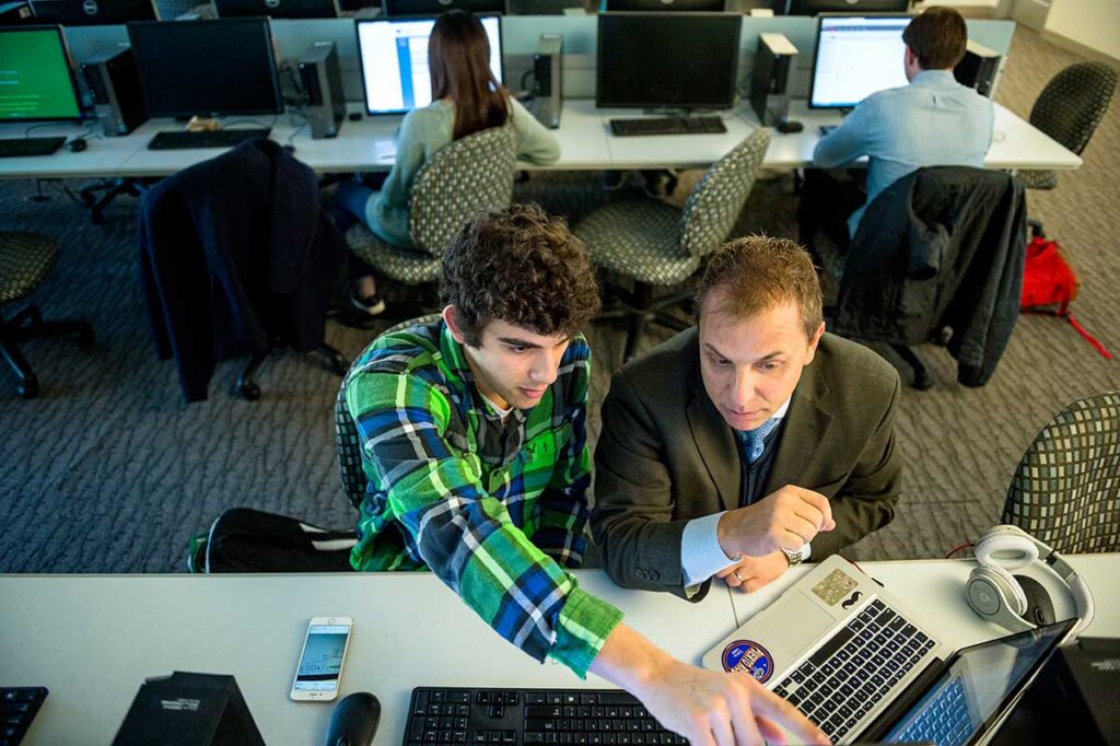 A Khoury student sitting at a desk points at his laptop while speaking with a faculty member