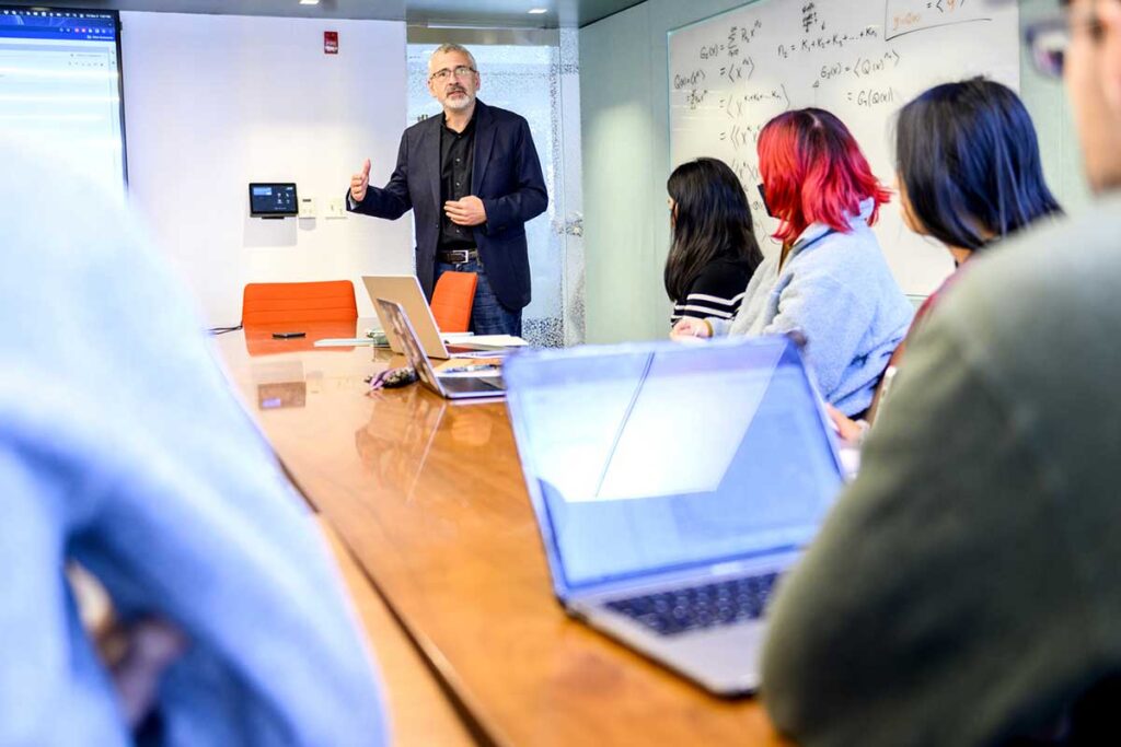 A Khoury professor stands at the head of a long table giving a presentation to several students seated around the tabl