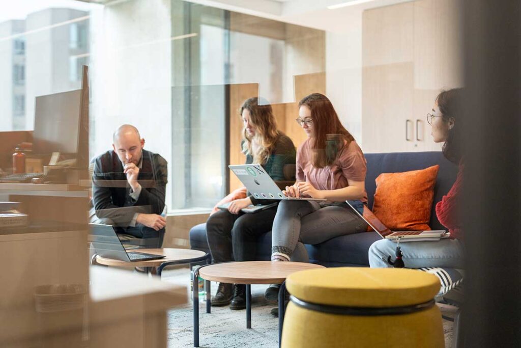 A Khoury faculty member sits in a lab while three students sit on a couch with their laptops open in front of them.