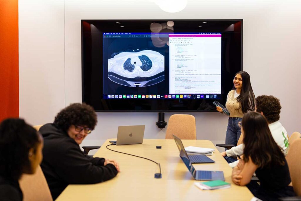 A student stands in front of a large display screen showing medical imaging while four students sit at a conference table.