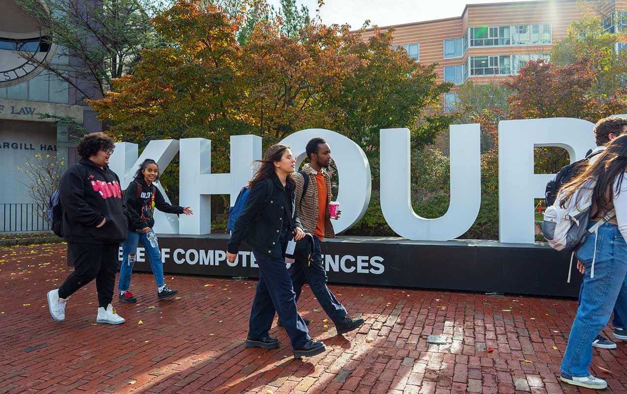 Four students walk on a brick sidewalk in front of a large, white Khoury College of Computer Sciences sign on the Boston campus