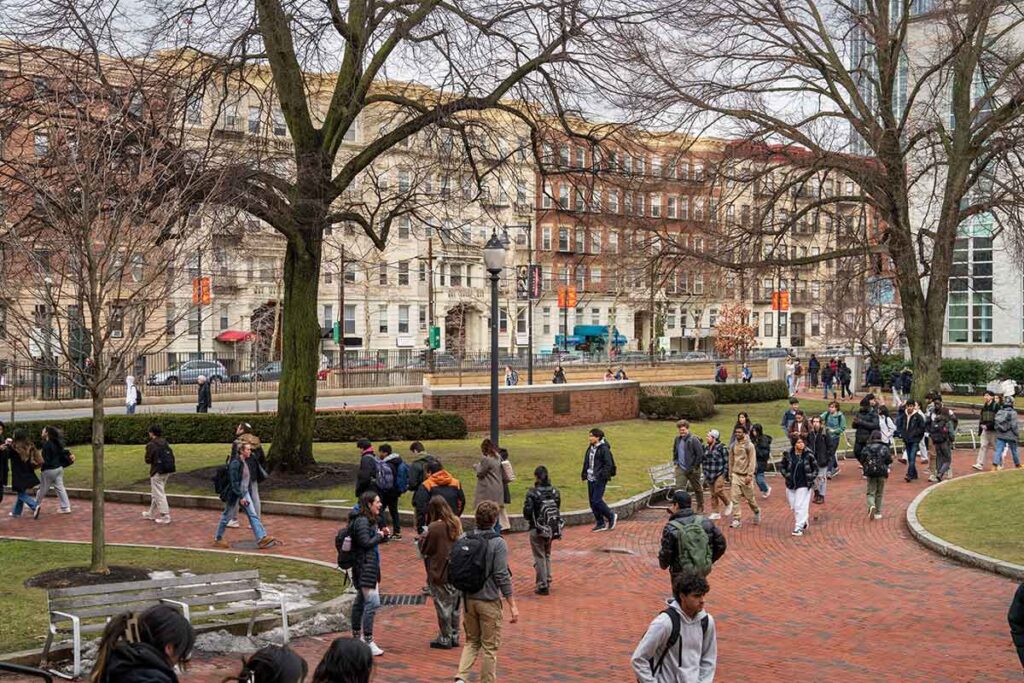 A wide view of Krentzman Quad at Northeastern's Boston campus. Dozens of students walk on brick sidewalks and Huntington Avenue and apartment buildings are visible in the background.