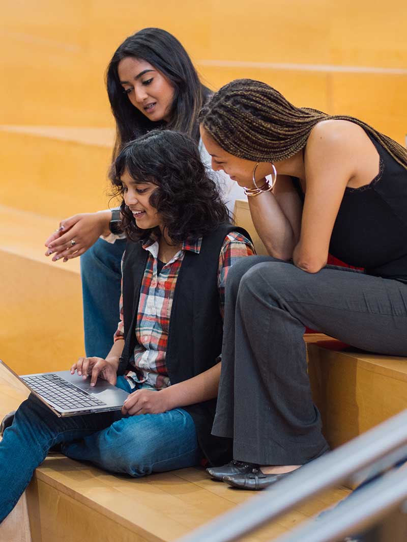 Three students sitting on wooden steps view a laptop