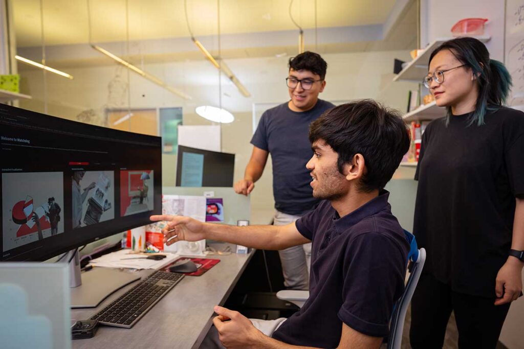 Three Khoury researchers look a a computer screen in a Khoury lab. One of the students is sitting in a chair pointing at the screen.