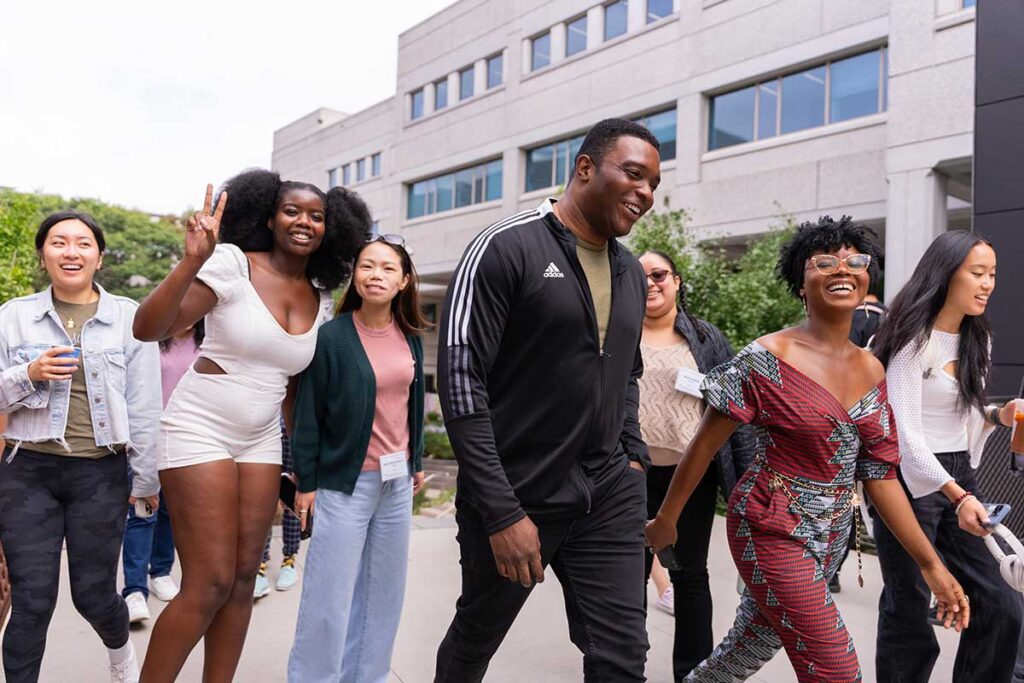 A group of 7 Northeastern students walk on a path on the Boston Campus with Snell Library in the background