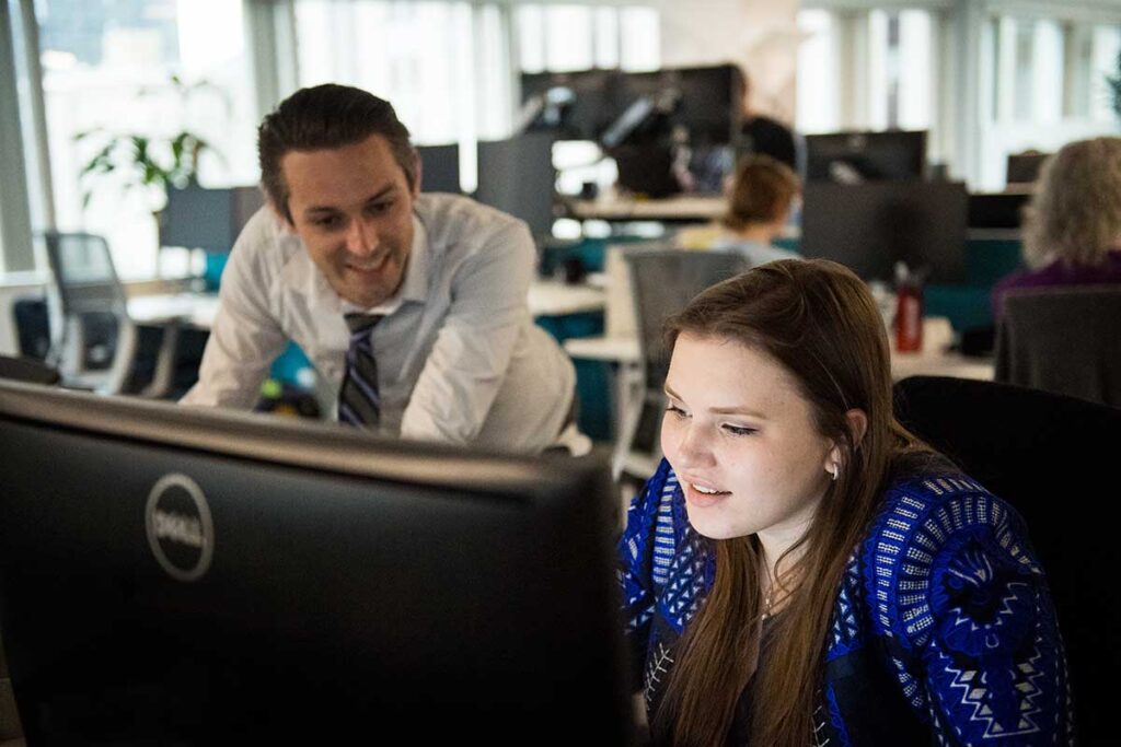 A student sits at a desk and does work at a computer in a Northeastern lab. A faculty member is leaning on the desk and viewing the screen.