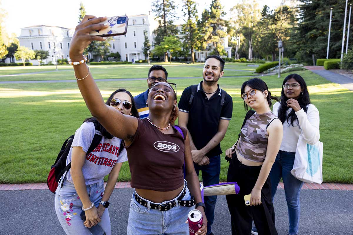 A student takes a selfie with five other students in the background in a courtyard on the Oakland campus